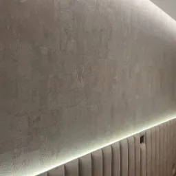 Ściana z beżową strukturą, oświetlona listwą LED, nad wezgłowiem łóżka z tapicerowanymi panelami w kolorze beżowym, widoczny fragment regału.