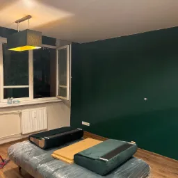 Świeżo pomalowana ściana w kolorze butelkowej zieleni w pokoju z podłogą z paneli, zabezpieczone meble pod folią, widoczny mop do podłogi.