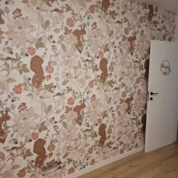 Ściana pokryta tapetą w kwiaty i wiewiórki, z widocznymi gniazdkami elektrycznymi i białą listwą przypodłogową oraz drzwiami z dekoracją.