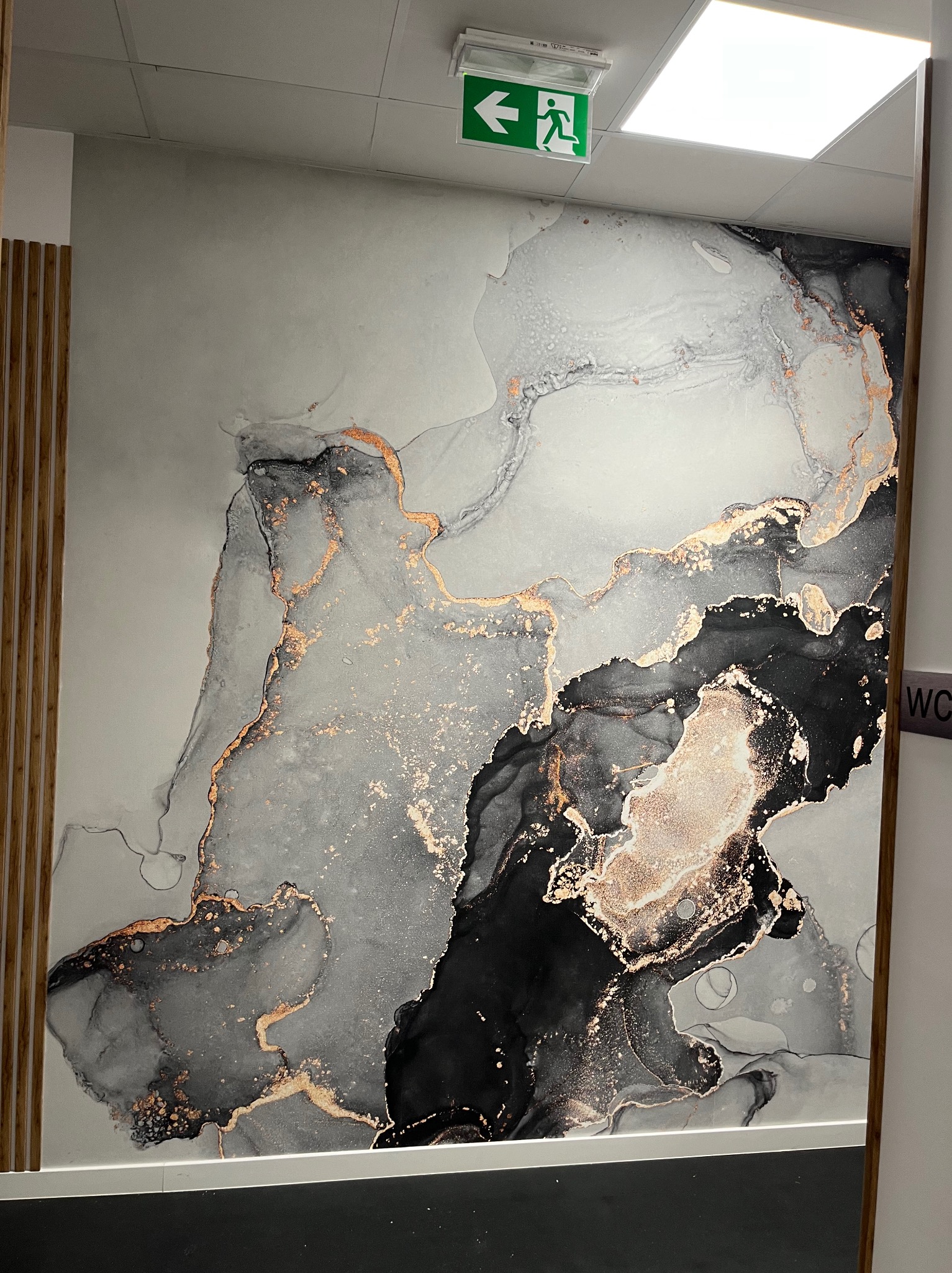 Nowoczesna fototapeta w odcieniach szarości, czerni i złota imitująca marmur, z widocznym fragmentem sufitu z lampą i znakiem ewakuacyjnym oraz elementami drewnianej boazerii po lewej stronie.