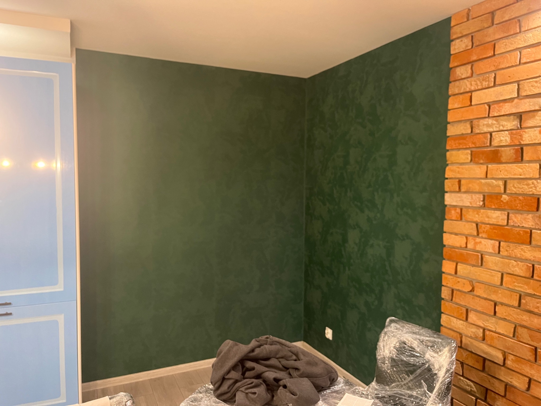 Pokój po remoncie: ściany w odcieniach zieleni z efektem dekoracyjnym, fragment ściany z cegły, zabezpieczone meble folią malarską.