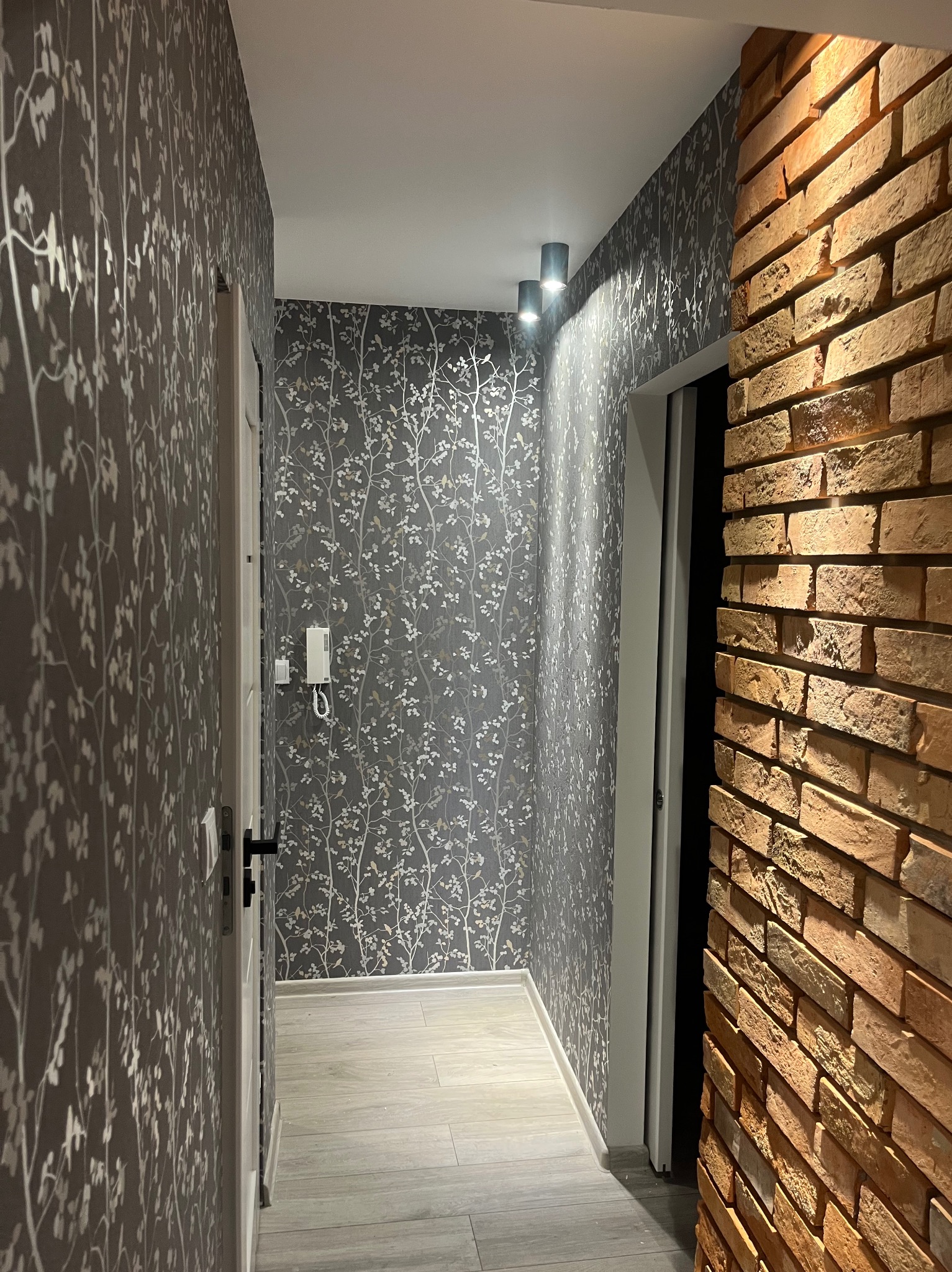 Korytarz z tapetą w odcieniach szarości z motywem drzew, oświetleniem punktowym i dekoracyjną ścianą z cegły.