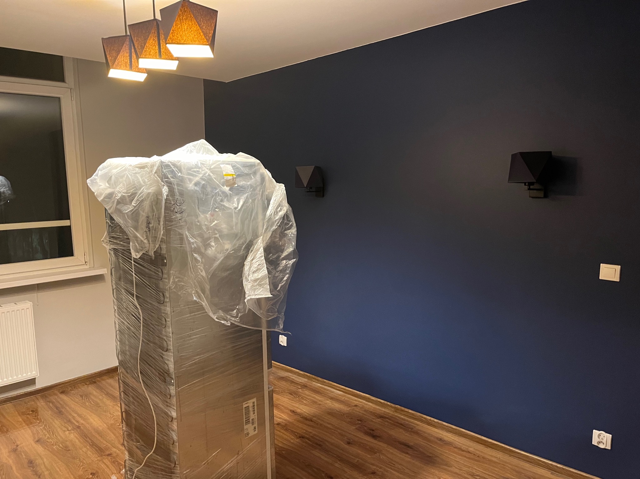 Pokój po odświeżeniu: ściana w odcieniach granatu z nowymi kinkietami, podłoga drewniana, mebel zabezpieczony folią ochronną przed zabrudzeniem.