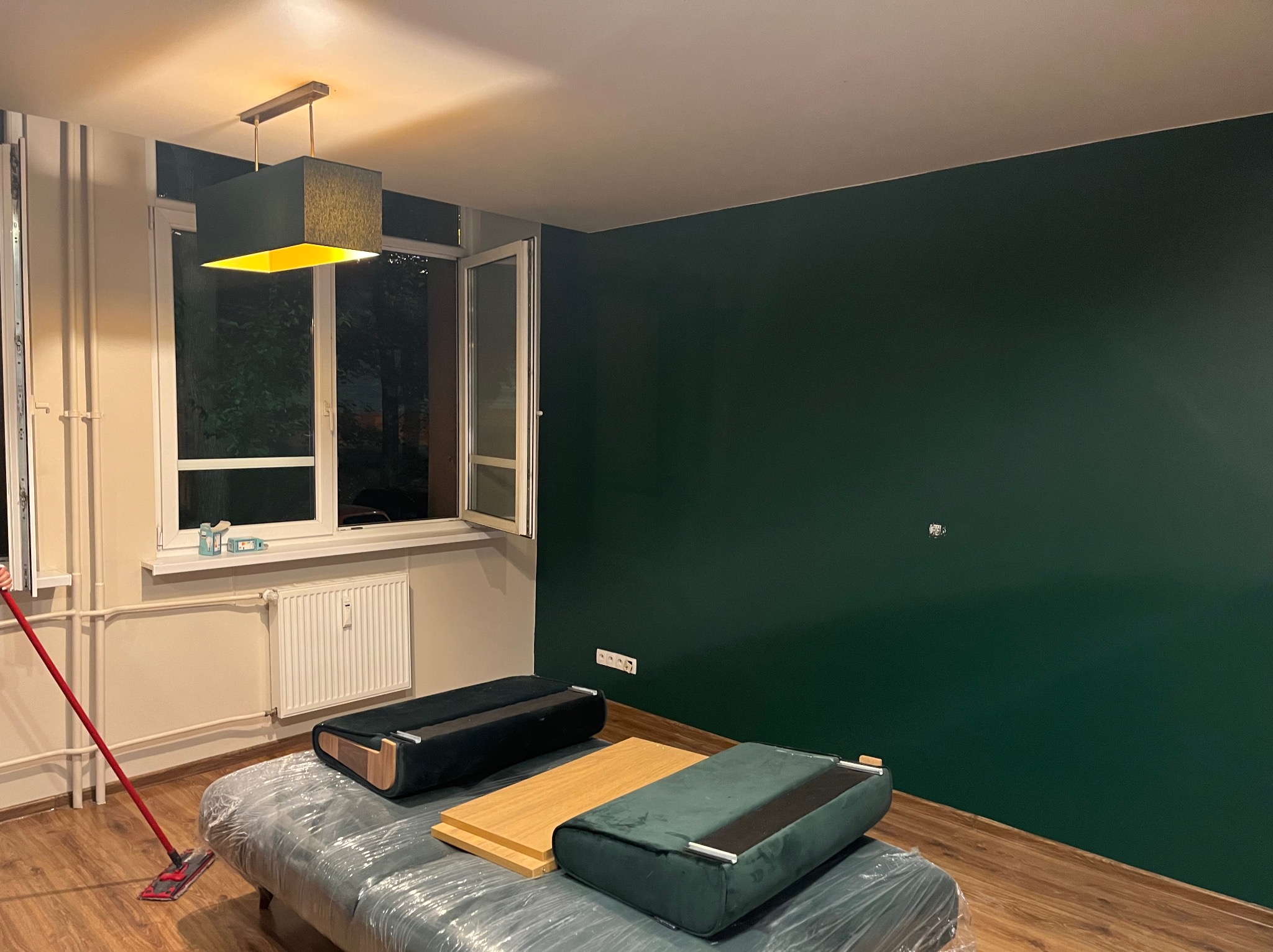 Świeżo pomalowana ściana w kolorze butelkowej zieleni w pokoju z podłogą z paneli, zabezpieczone meble pod folią, widoczny mop do podłogi.