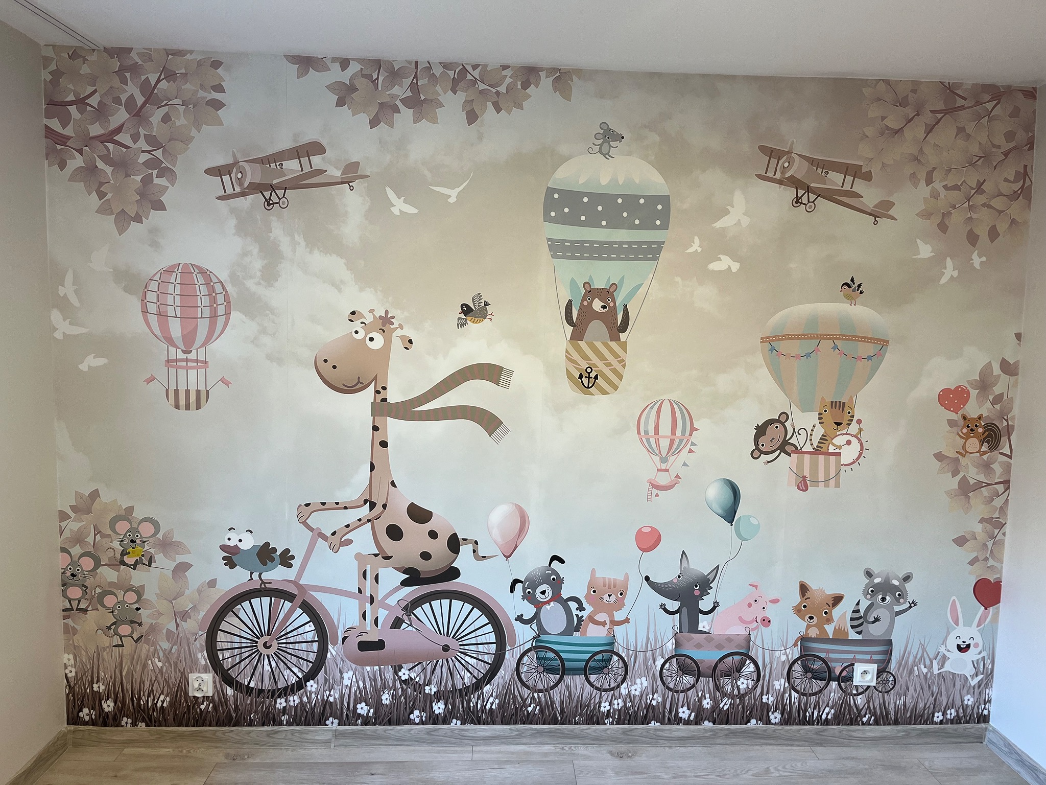 Dziecięca tapeta na ścianie z motywem żyrafy na rowerze ciągnącej wagoniki z zwierzętami, balonów i samolotów w pastelowych kolorach.