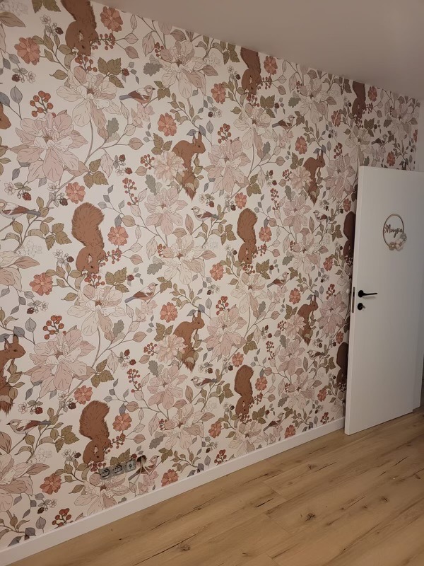 Ściana pokryta tapetą w kwiaty i wiewiórki, z widocznymi gniazdkami elektrycznymi i białą listwą przypodłogową oraz drzwiami z dekoracją.