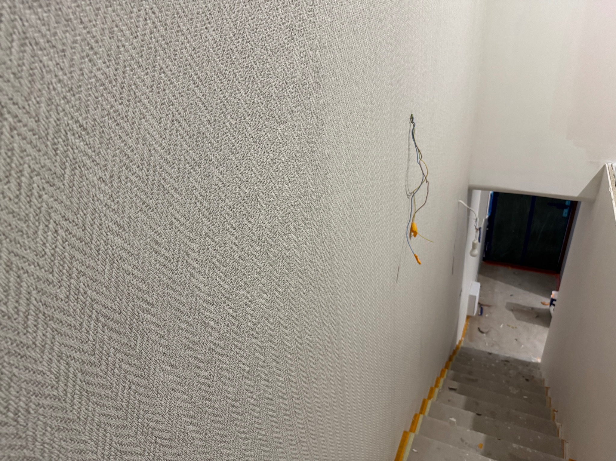 Ściana w klatce schodowej pokryta szarą tapetą w delikatny wzór jodełki, widoczne wystające przewody elektryczne i fragment schodów z zabezpieczeniami.