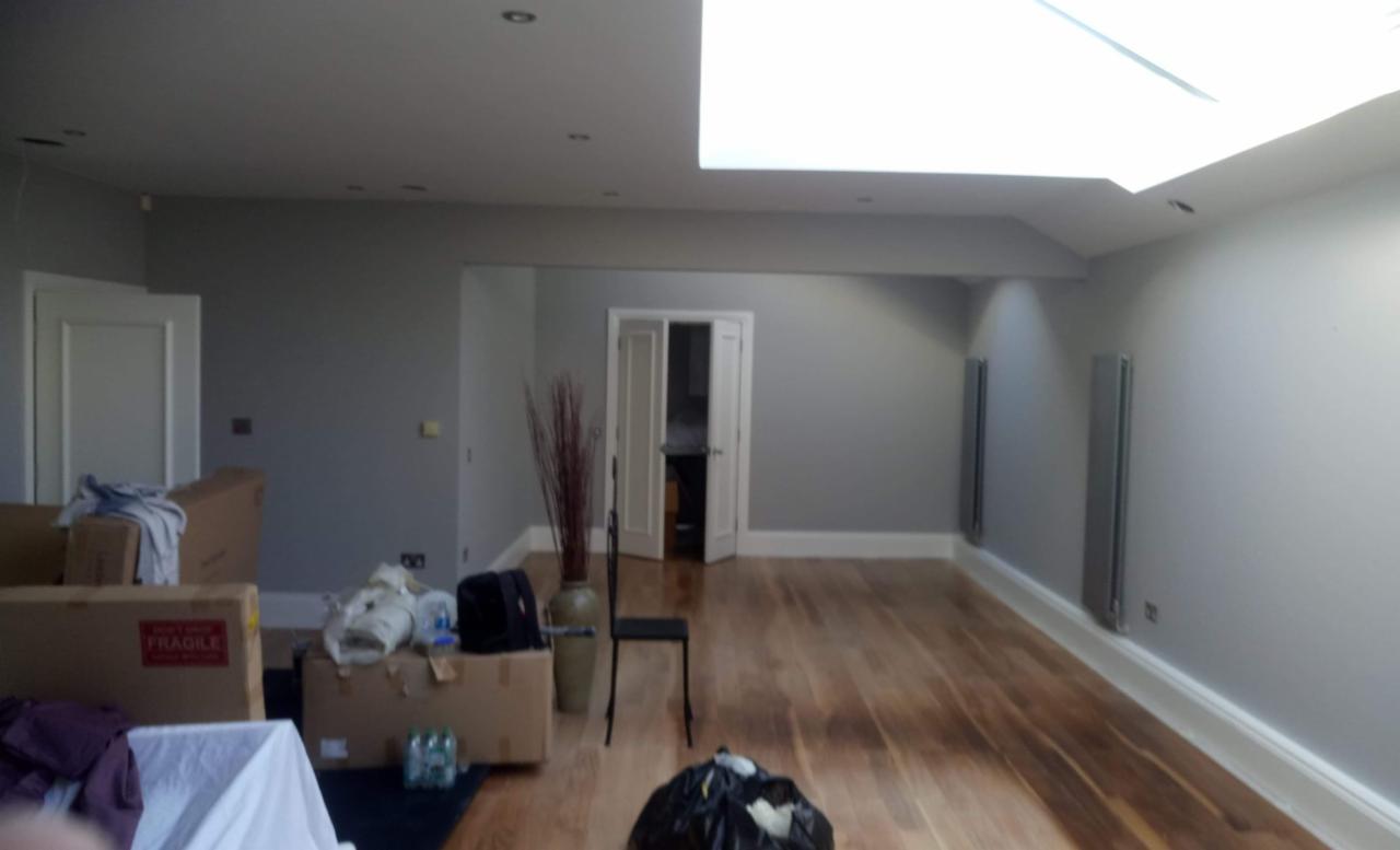 Wnętrze pokoju po malowaniu ścian na szaro, widoczne kartony z rzeczami, drewniana podłoga, grzejniki panelowe, białe listwy przypodłogowe i otwarte drzwi do innego pomieszczenia.