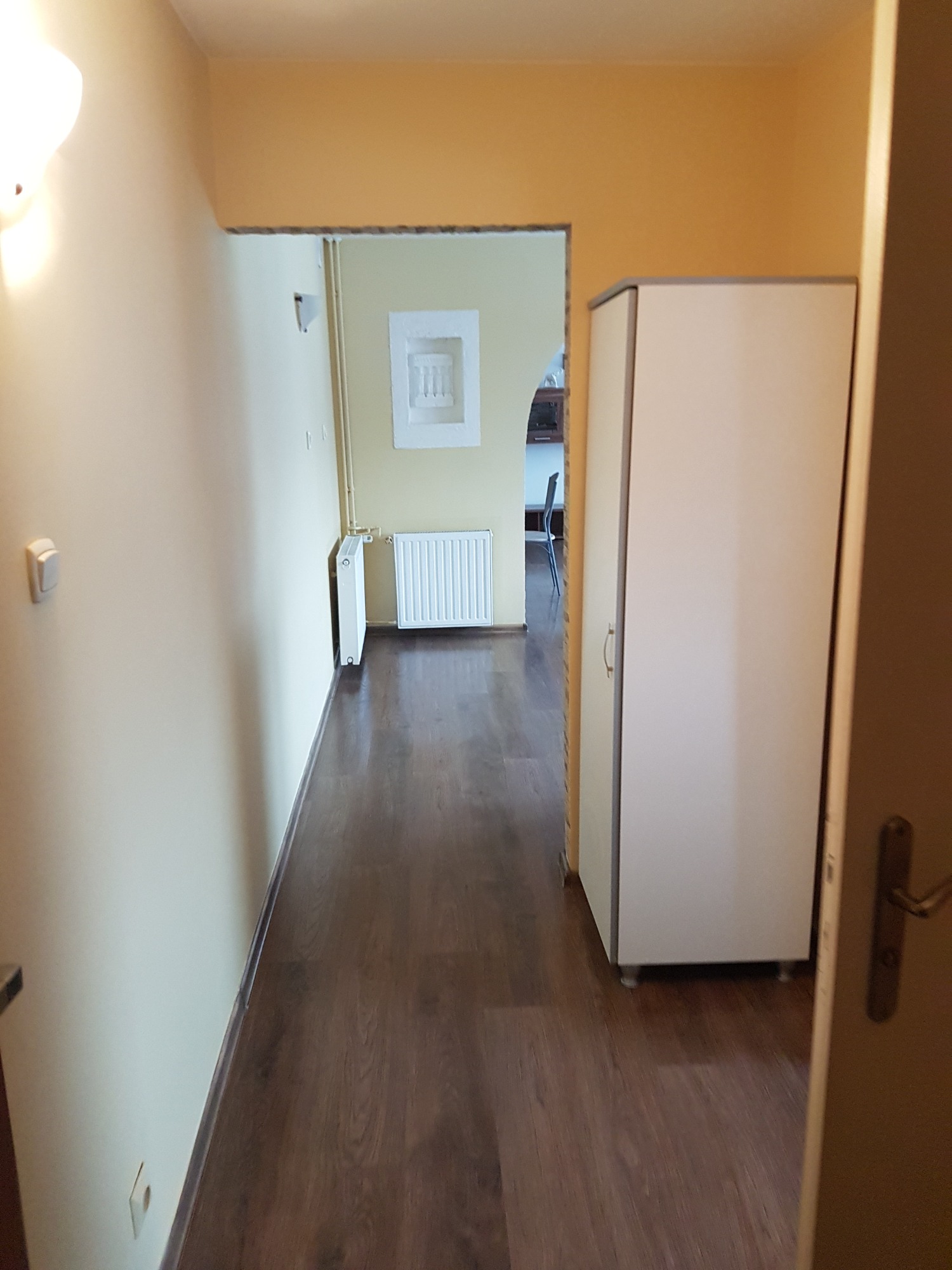 Korytarz w mieszkaniu po remoncie z pomalowanymi ścianami na odcienie żółci i brązu, widoczna szafa i grzejnik, podłoga wyłożona panelami imitującymi drewno.