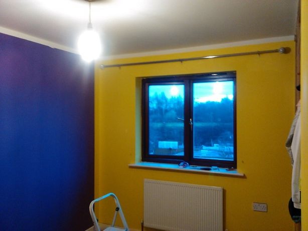 Pokój po malowaniu: jedna ściana w kolorze głębokiego fioletu, druga jaskrawożółta, okno z widokiem na zieleń, grzejnik pod oknem, drabinka malarska w rogu.