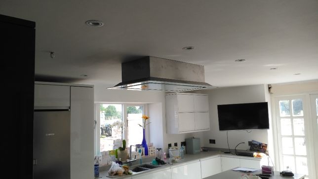 Nowoczesna kuchnia z białymi szafkami, stalową lodówką i okapem kuchennym nad wyspą, widok na okno z roślinami i kwiatami.