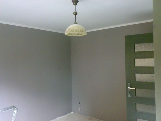 Pokój po malowaniu na szaro, widoczna nowa lampa sufitowa z wzorem meandrycznym, biała listwa przysufitowa i zielone drzwi z przeszkleniami.