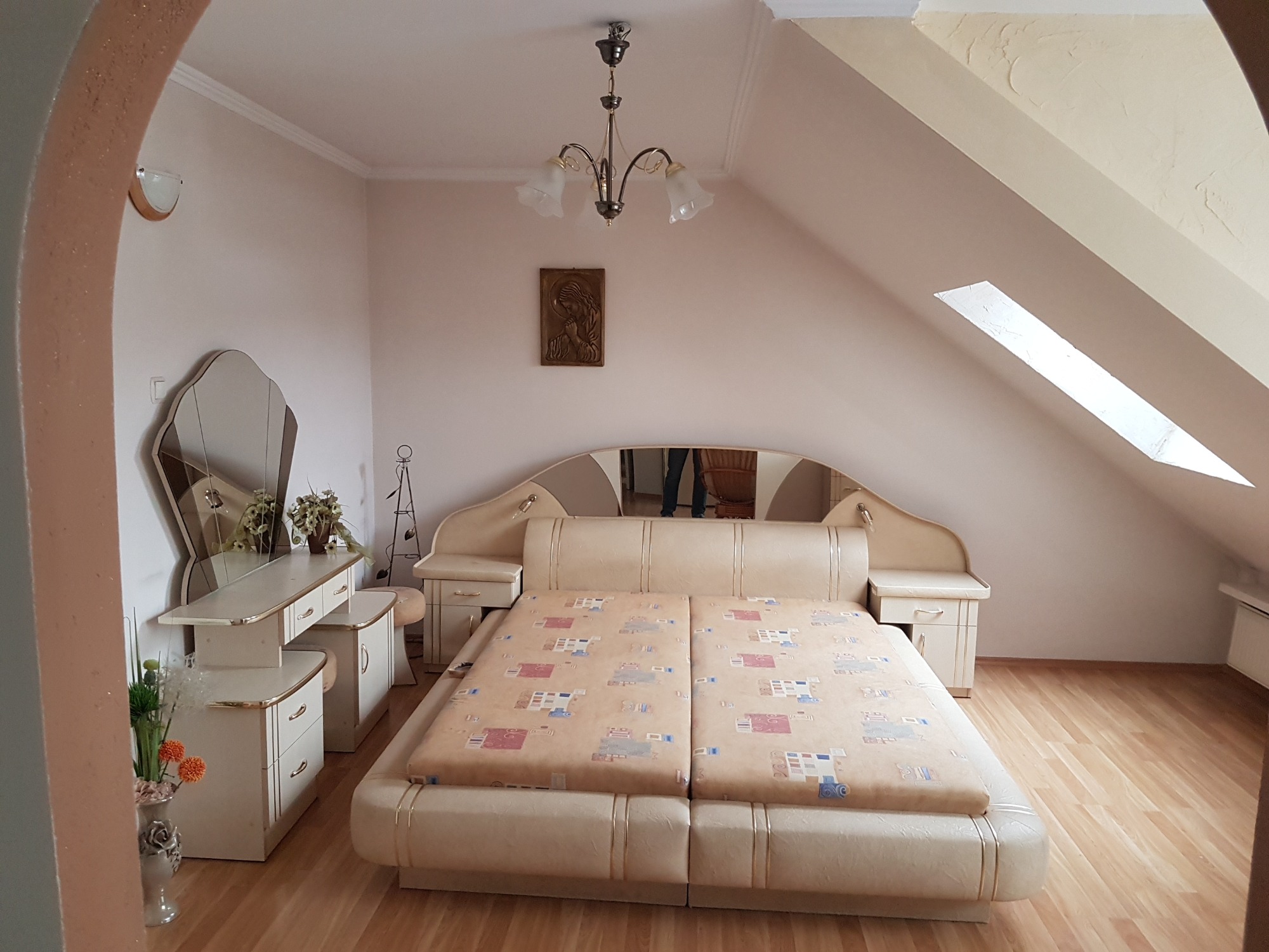 Jasna sypialnia na poddaszu z łóżkiem w stylu glamour, toaletką z lustrem i drewnianą podłogą, oświetlona żyrandolem.
