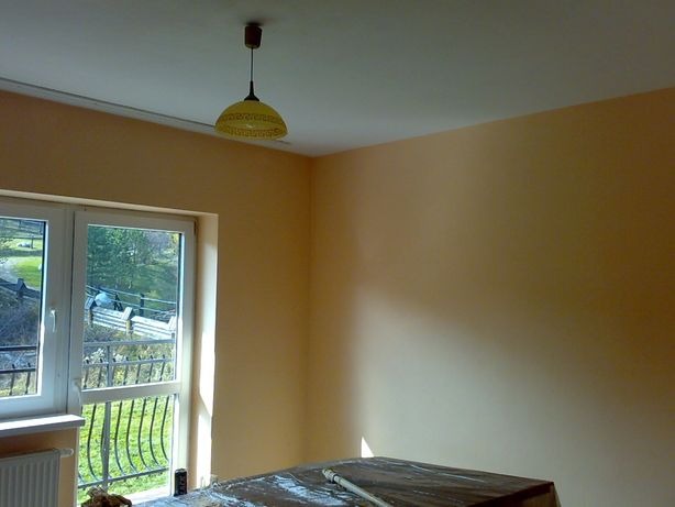 Pomarańczowe ściany w pokoju po malowaniu, widoczna lampa z żółtym abażurem i okno z widokiem na zewnątrz, meble przykryte folią ochronną.