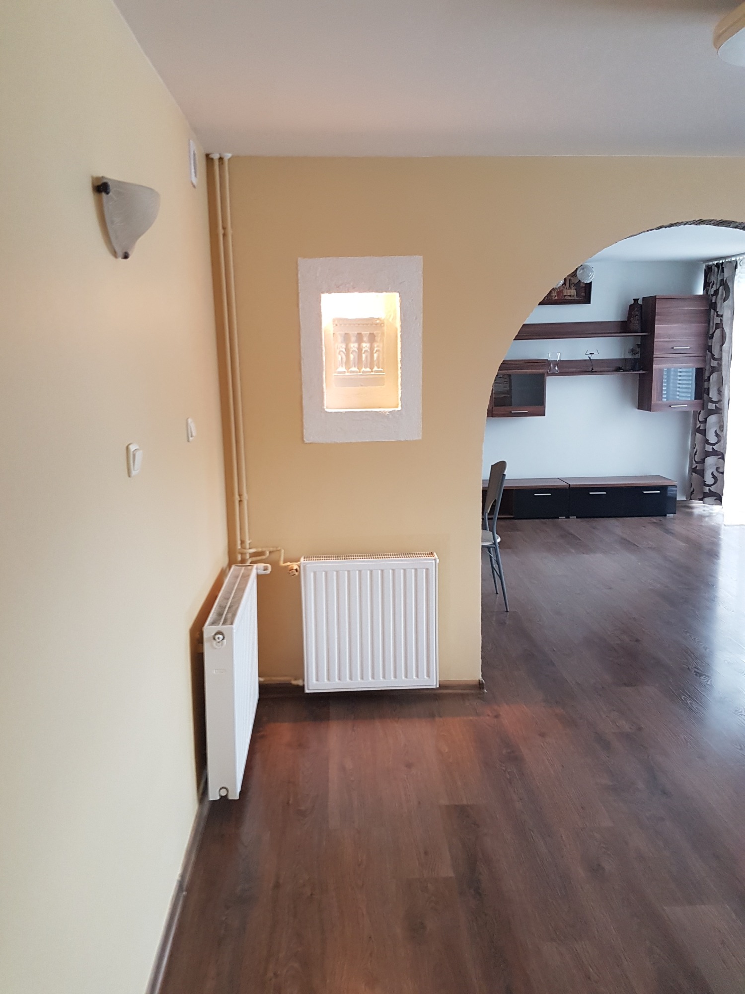 Salon z kremowymi ścianami, białymi grzejnikami, drewnianą podłogą i dekoracyjnym oknem w ścianie, widok na umeblowany pokój przez łuk.