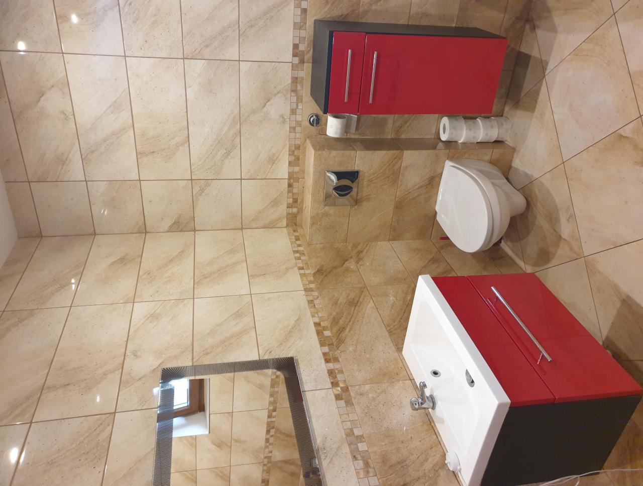 Wykończona łazienka z beżowymi płytkami, czerwono-czarnymi szafkami, białą umywalką i toaletą, widziana z góry pod kątem.