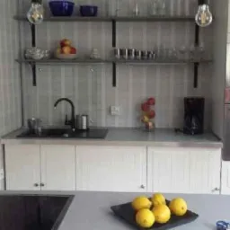 Jasna kuchnia z białymi szafkami, czarnym zlewem i baterią, otwartymi półkami z dekoracjami, piekarnikiem w zabudowie i talerzem cytryn na blacie.