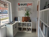 Jasne wnętrze biura EuroPVC z oknem PCV, białymi regałami i rośliną doniczkową. Na ścianie widoczny plakat informacyjny firmy. Ujęcie z perspektywy wejścia.