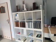 Ekspozycja próbek materiałów wykończeniowych na białym regale. Widoczne próbki oklein, blatów i innych materiałów w różnych kolorach i fakturach. Białe drzwi w tle.