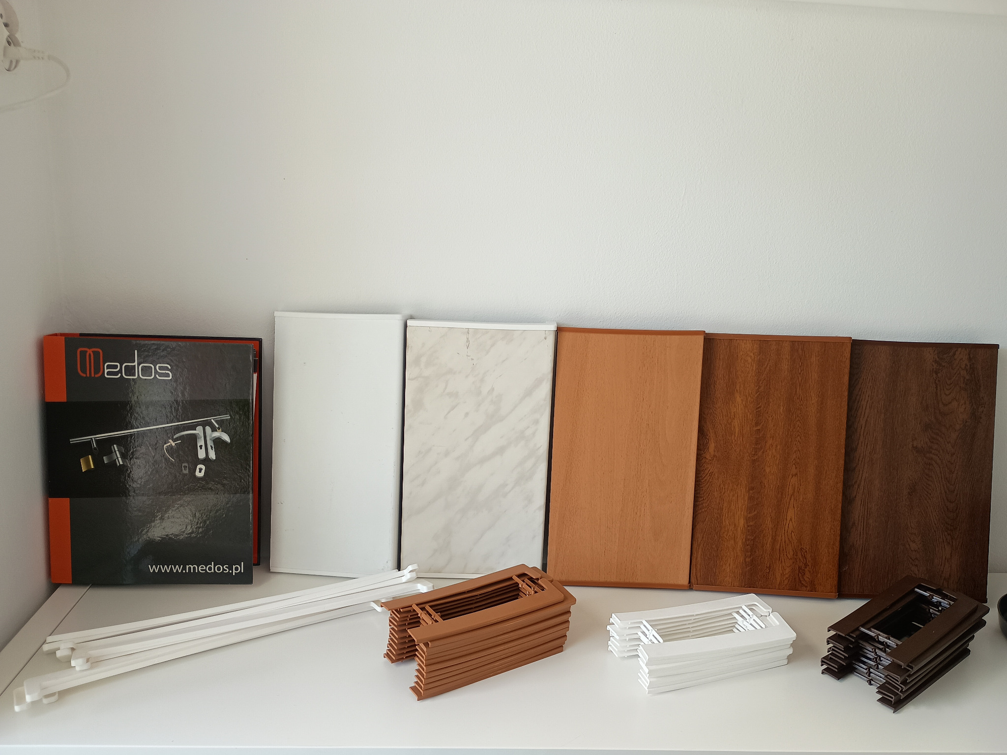 Próbki kolorów okiennych i profili w odcieniach bieli, marmuru i drewna, ułożone na białym blacie. Widoczna paczka z akcesoriami okiennymi.