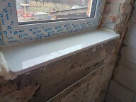 Biały parapet z PCV, zamontowany w surowym otworze okiennym z widoczną pianką montażową i fragmentami cegieł. Nowa instalacja, detale wykończenia.