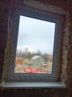 Białe okno PCV z widokiem na szarą, pochmurną scenerię ogrodu i szklarnię, osadzone w surowej, nieotynkowanej ścianie z cegieł.