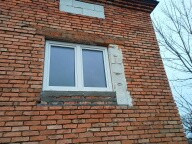 Białe okno PCV w surowej, ceglanej ścianie budynku w trakcie remontu, z widocznymi śladami prac murarskich i częściowym ociepleniem.  Ujęcie z dołu.