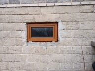 Małe, brązowe okno w surowej, jasnej ścianie z pustaków. Widoczne ślady po montażu, szara fuga. Minimalistyczny styl, proste wykończenie.