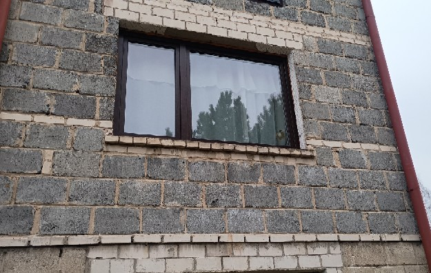 Brązowe okno PCV w szarym murze z betonu komórkowego, widok z zewnątrz. W szybie odbijają się drzewa i niebo. Czerwona rynna po prawej stronie.