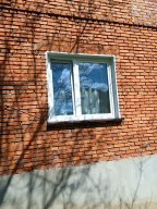 Białe okno PCV osadzone w ścianie z czerwonej cegły, z widocznym odbiciem błękitnego nieba i chmur. Cień drzewa na murze. Ujęcie z dołu.