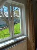 Białe okno PCV z widokiem na zielony ogród i budynek. Zasłona z geometrycznym wzorem po prawej stronie. Grzejnik pod oknem. Jasne wnętrze.