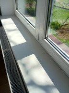 Białe okno PCV z parapetem z konglomeratu w neutralnym świetle dziennym. Widoczny fragment grzejnika. Ujęcie z bliska, ukazuje detale wykończenia.