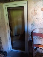 Białe, wąskie okno PCV w surowej, starej ścianie z widoczną fakturą i elementami drewnianymi. Kątowe ujęcie z krzesłem w kadrze, wnętrze słabo widoczne.