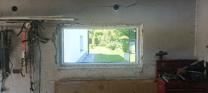 Montaż okna PCV w surowym pomieszczeniu. Widok na ogród przez nowe okno. Na ścianach widoczne ślady po pracach budowlanych, narzędzia i akcesoria.