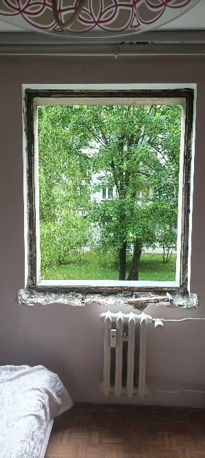 Demontaż starego okna, widok na zieleń za oknem. Widoczne ślady po starej ramie okiennej, grzejnik pod oknem i fragment pokoju z łóżkiem.
