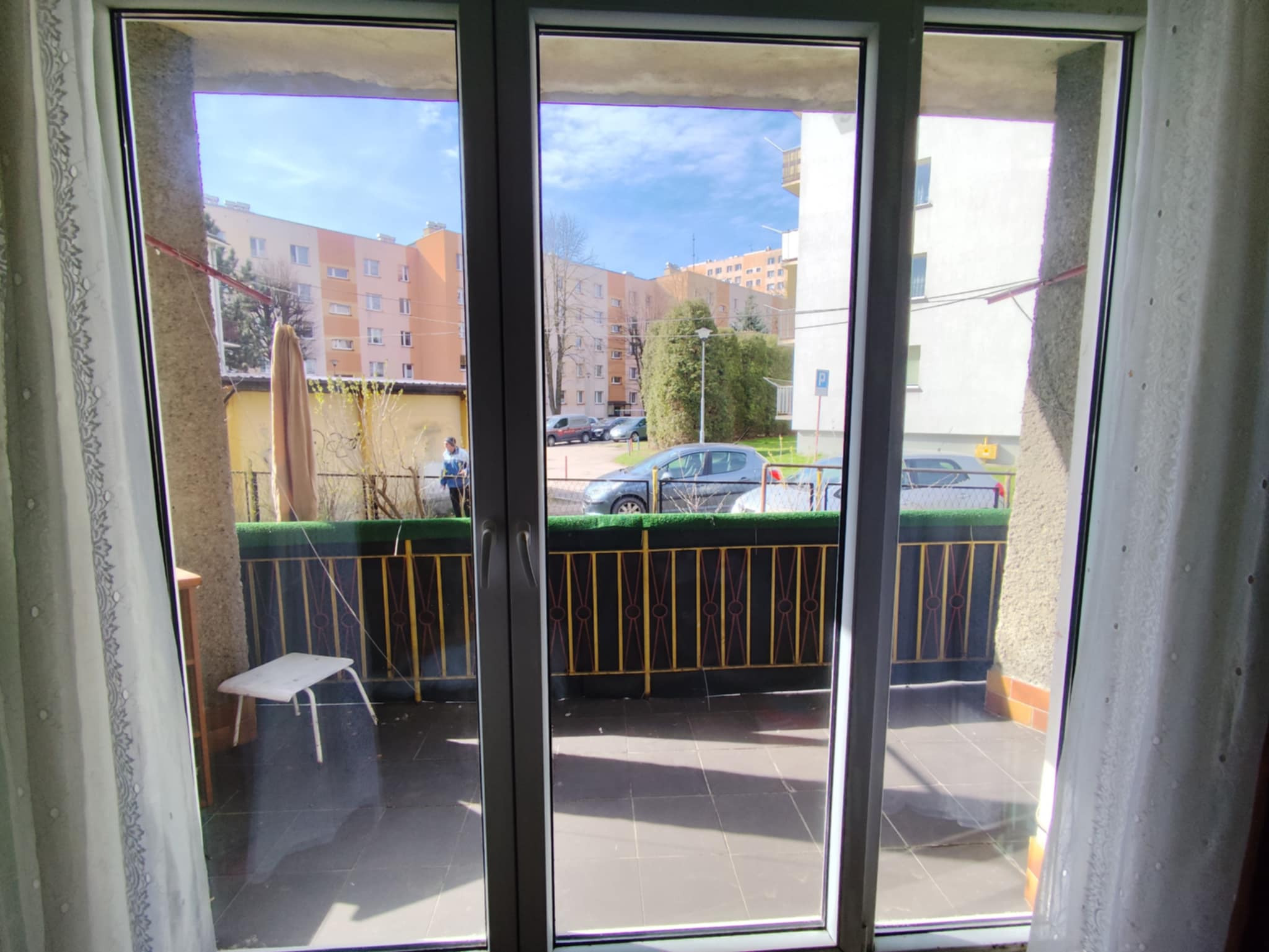 Białe, rozsuwane okno PCV z widokiem na balkon z czarną podłogą i metalową barierką. W tle budynki mieszkalne i osoba.