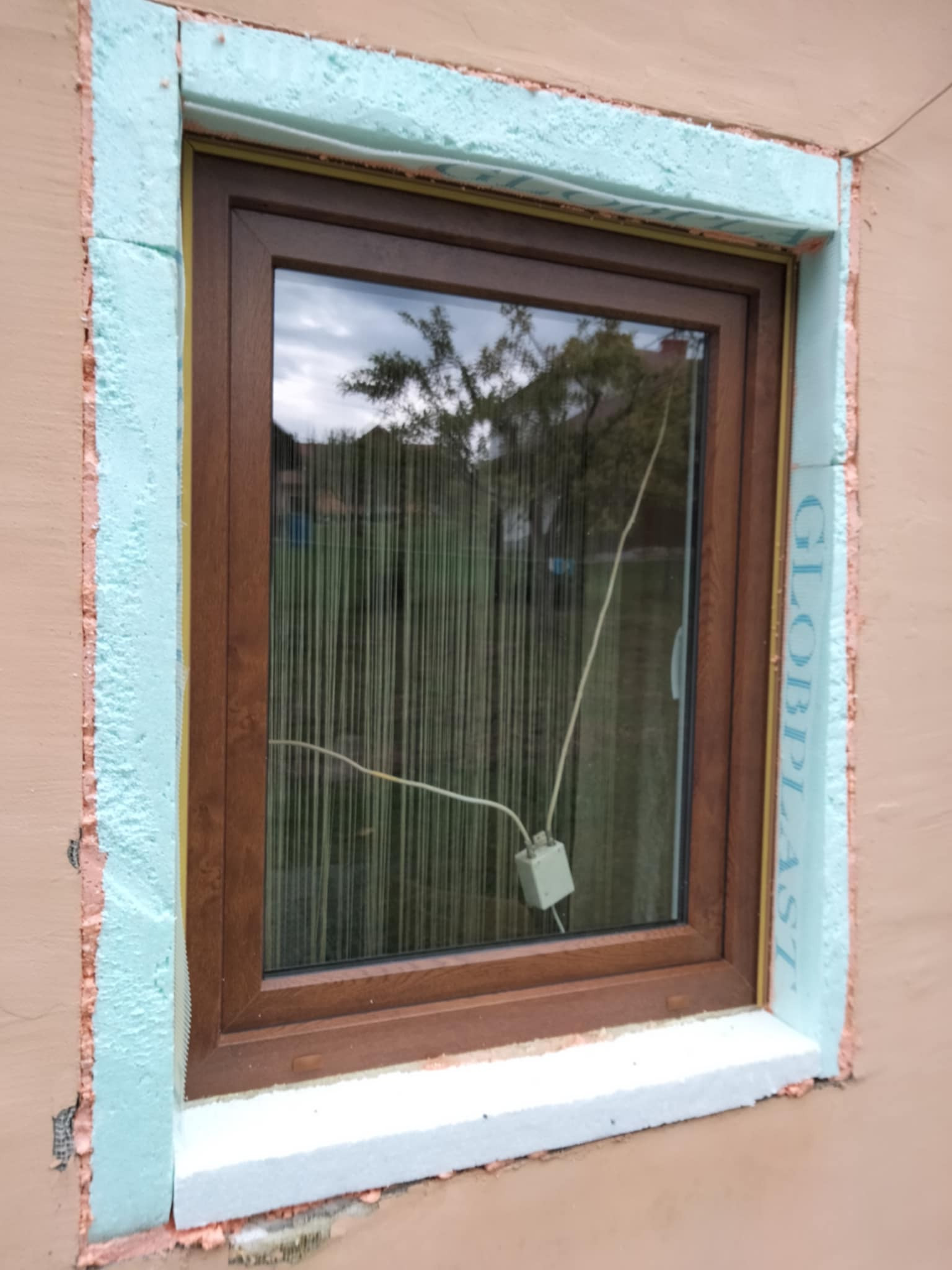 Montaż okna z brązową ramą i niebieskim ociepleniem. Widoczne elementy mocujące i odbicie drzew w szybie. Częściowo otynkowana ściana zewnętrzna.