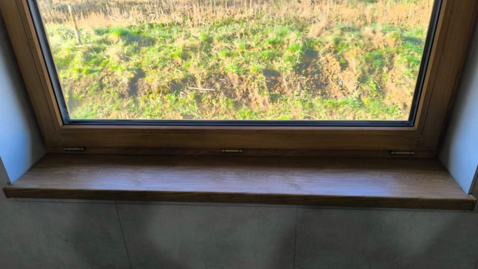 Drewniane okno dachowe z parapetem, widok na zewnątrz. Jasne wykończenie ramy i parapetu, naturalne światło, tło z zielenią i ziemią. Detal montażu okna.