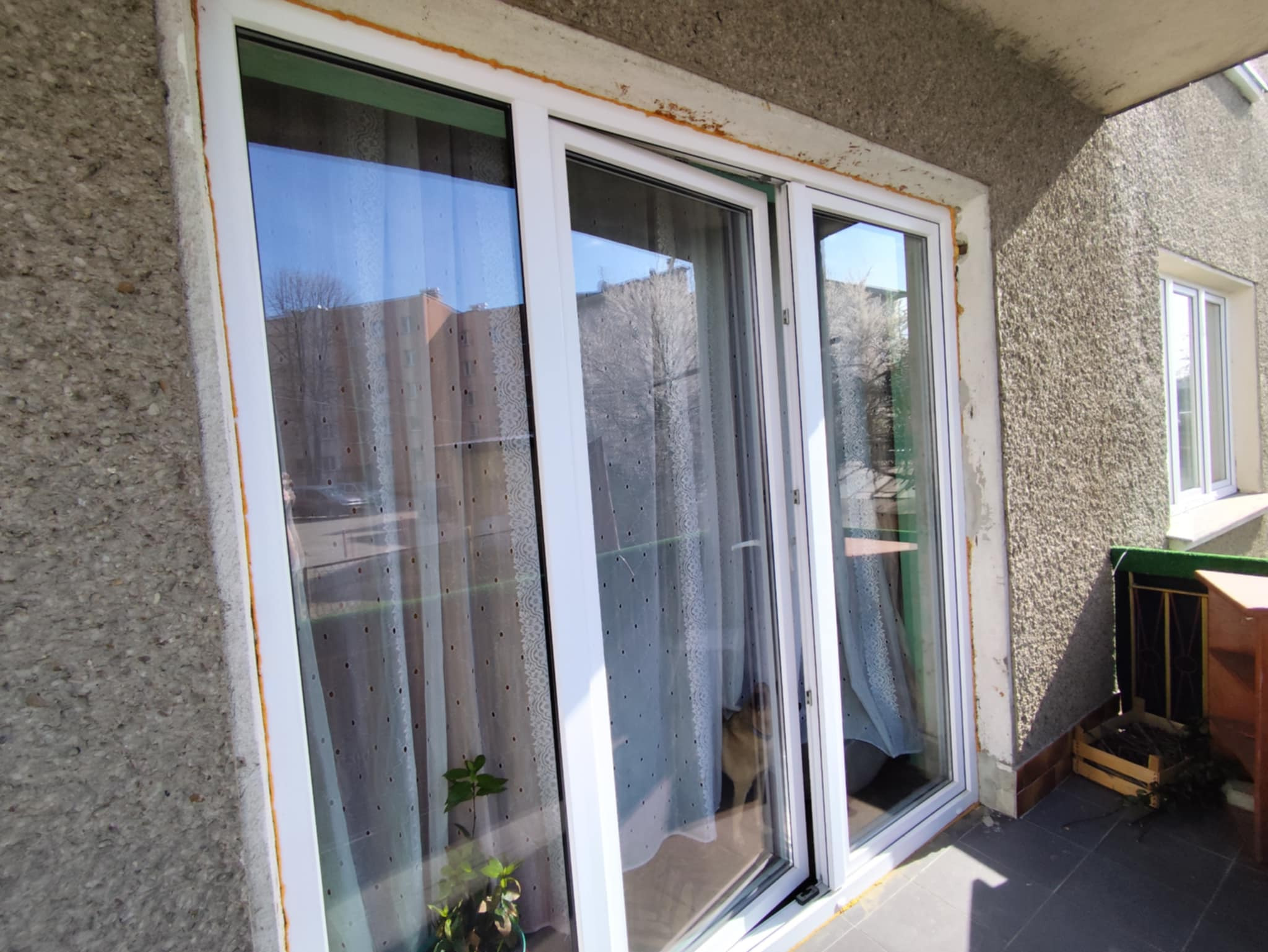 Balkon z białymi drzwiami przesuwnymi, widoczne ślady uszczelniania. W tle budynek mieszkalny odbija się w szybach. Na balkonie roślina w doniczce i skrzynka z gałęziami.