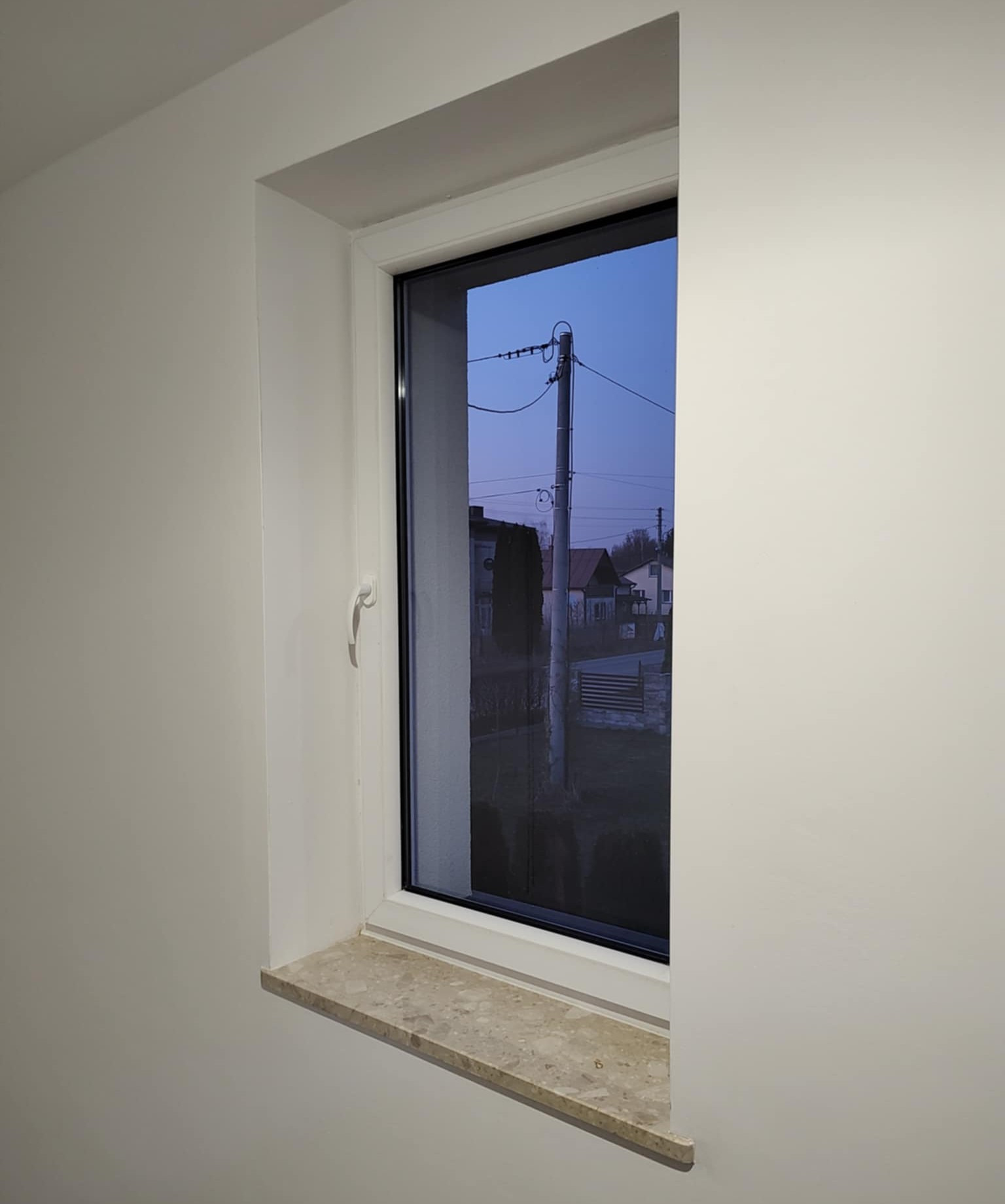 Białe okno PCV z marmurowym parapetem w jasnym wnętrzu, widok na zewnątrz na słup elektryczny i domy o zmierzchu. Minimalistyczny design.