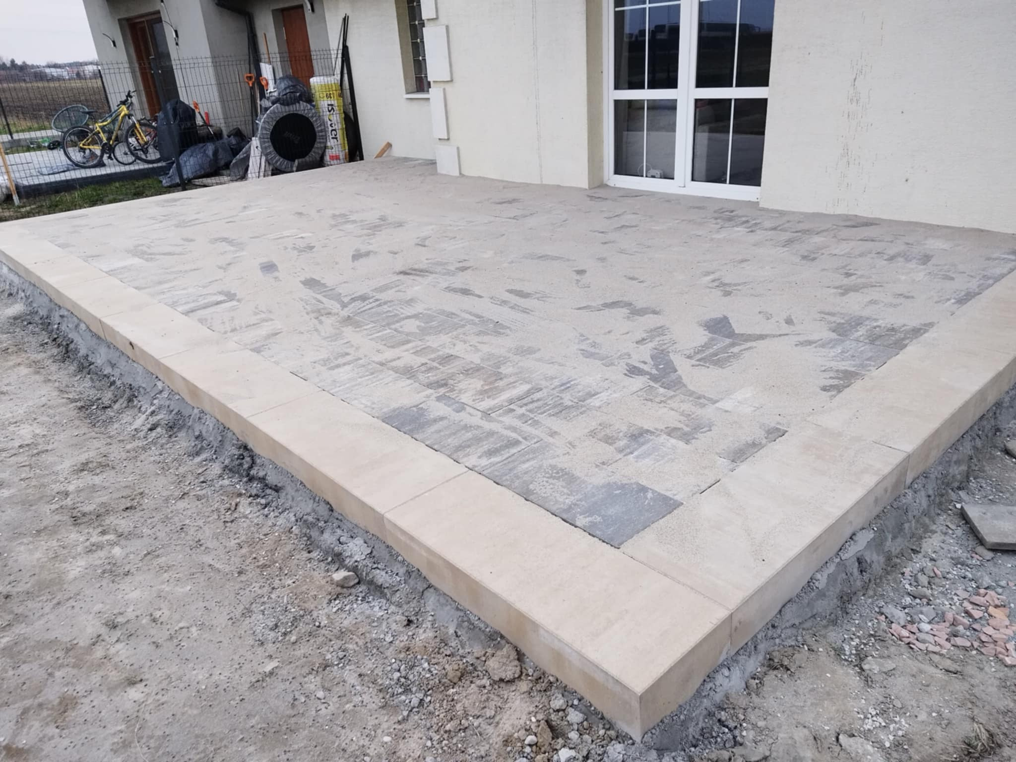 Wykończony taras z kostki brukowej w odcieniach szarości i beżu, obrzeża z jasnych płyt. Widok na dom z białymi drzwiami tarasowymi i elementy ogrodu w tle.