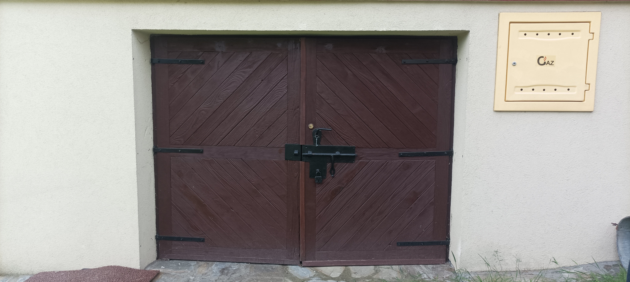 Dwuskrzydłowe, brązowe drzwi zewnętrzne z ukośnym wzorem desek i czarnym zamkiem, na tle beżowej ściany z żółtą skrzynką gazową. Widoczne elementy otoczenia.