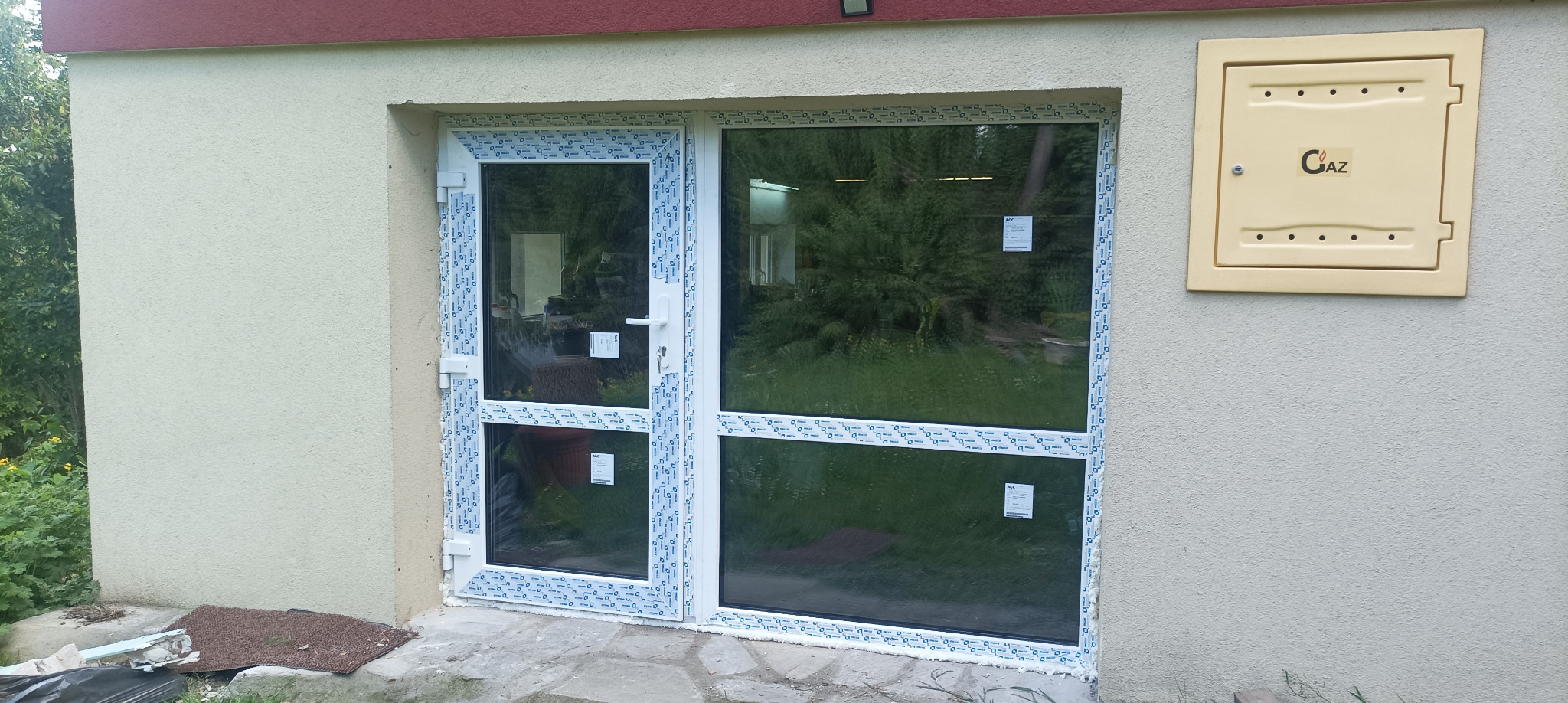 Białe drzwi PCV z dużym przeszkleniem, zamontowane w elewacji budynku. Obok skrzynka gazowa. Widoczne ślady montażu i otoczenie budynku.