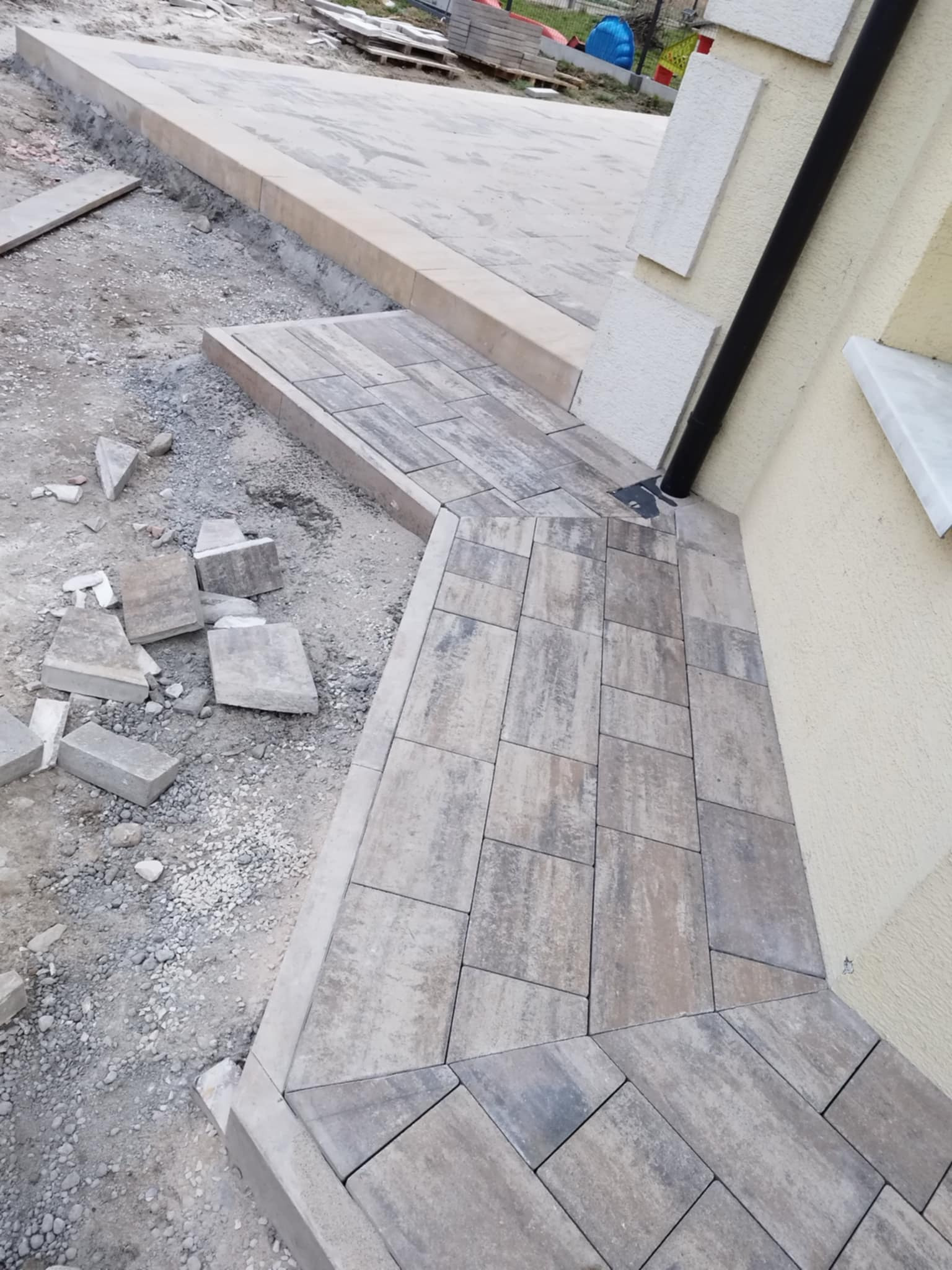 Ułożona kostka brukowa przy ścianie budynku z rurą spustową, widoczne luźne kostki i przygotowane podłoże. Kątowe ułożenie kostki przy ścianie.