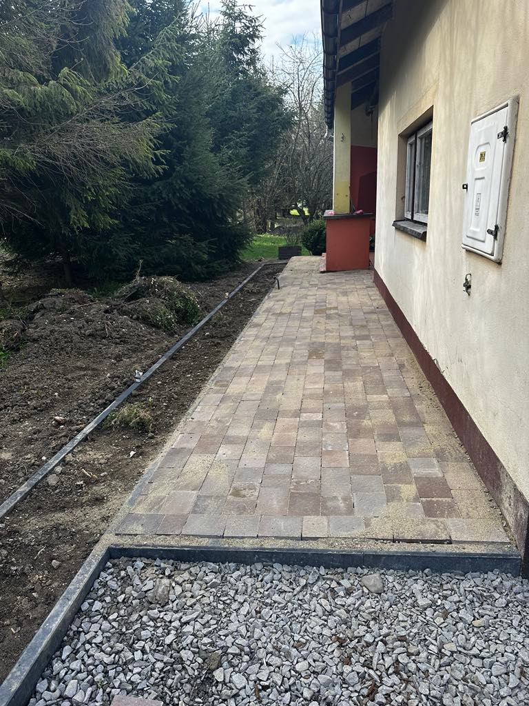 Ułożona kostka brukowa tworząca ścieżkę wzdłuż budynku, obrzeża z kamienia. Widoczne przygotowane podłoże pod dalsze prace ziemne. Ujęcie z niskiej perspektywy.