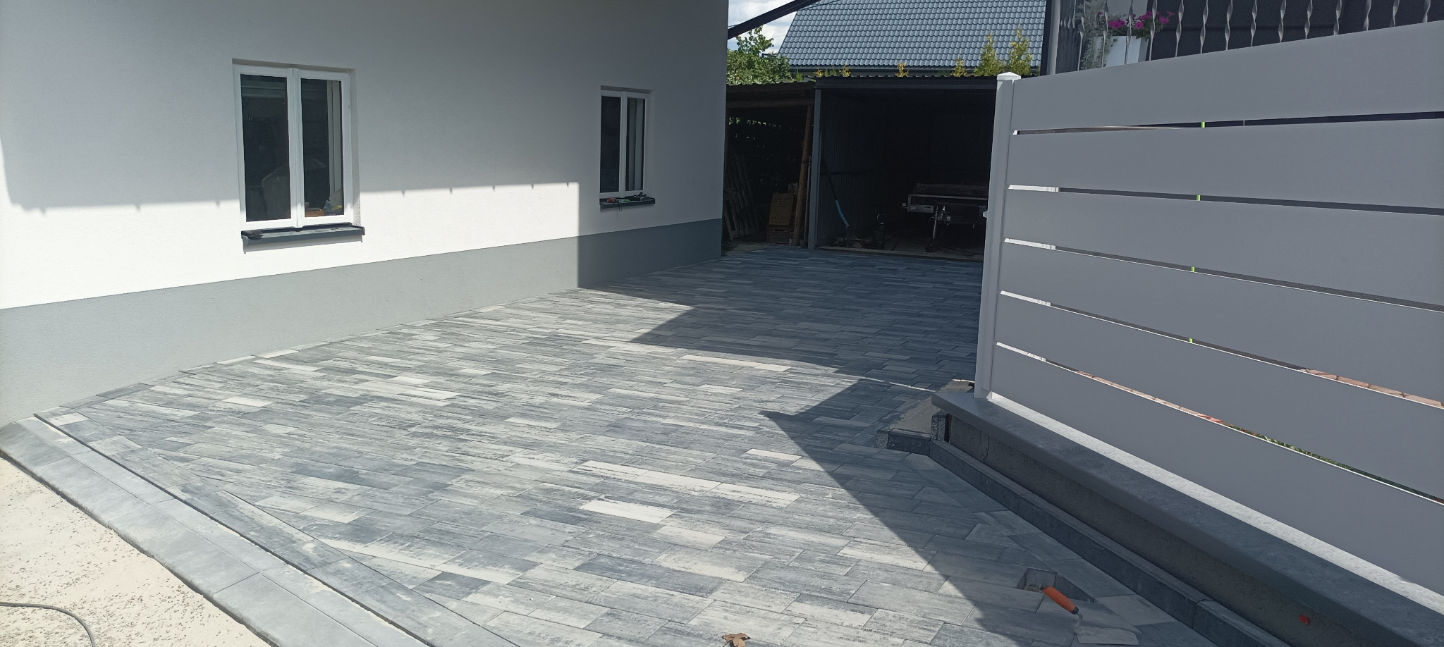 Ułożona kostka brukowa w szaro-niebieskich odcieniach na podjeździe, widok z perspektywy bramy wjazdowej. W tle biały budynek z oknami i garaż.