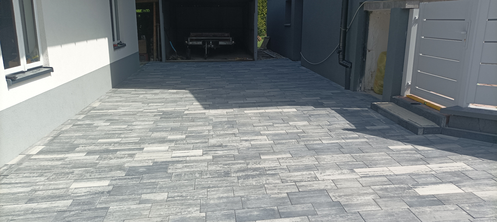 Ułożona kostka brukowa w odcieniach szarości, prowadząca do garażu i furtki. Widoczne detale: wzór kostki imitujący drewno, cień padający na powierzchnię.