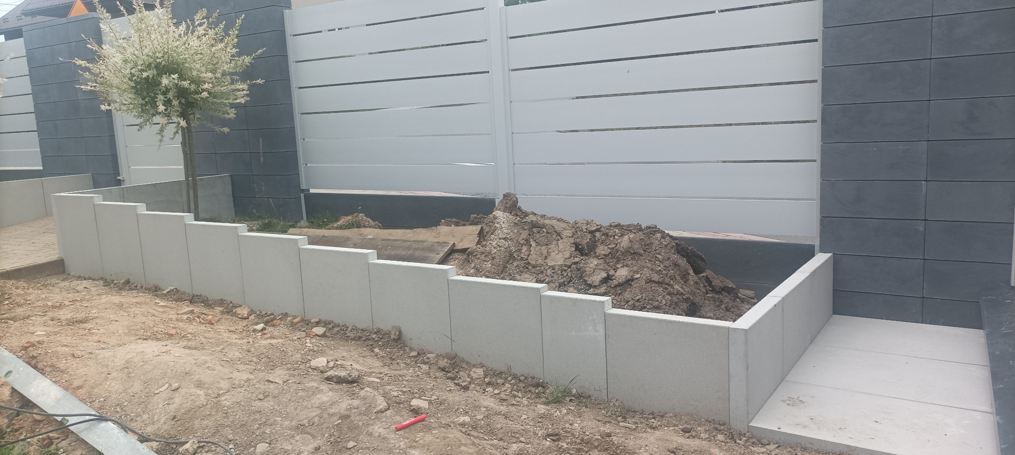Nowoczesne ogrodzenie z białymi przęsłami i szarymi słupkami z bloczków betonowych. W tle drzewo. Na pierwszym planie podmurówka z bloczków i ziemia.