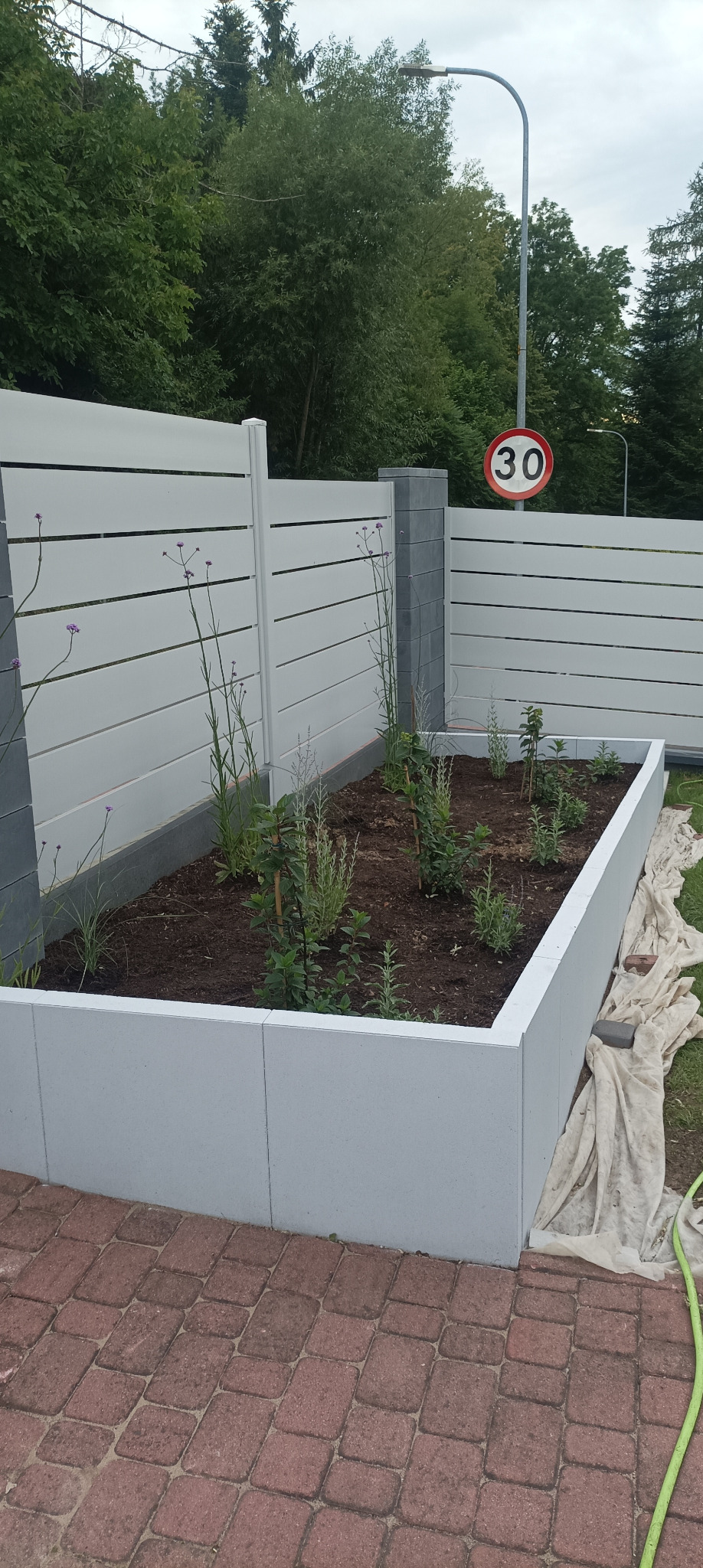 Nowoczesny, biały płot panelowy z betonowymi słupkami i podwyższona rabata z roślinami. W tle znak drogowy ograniczenia prędkości do 30 km/h.