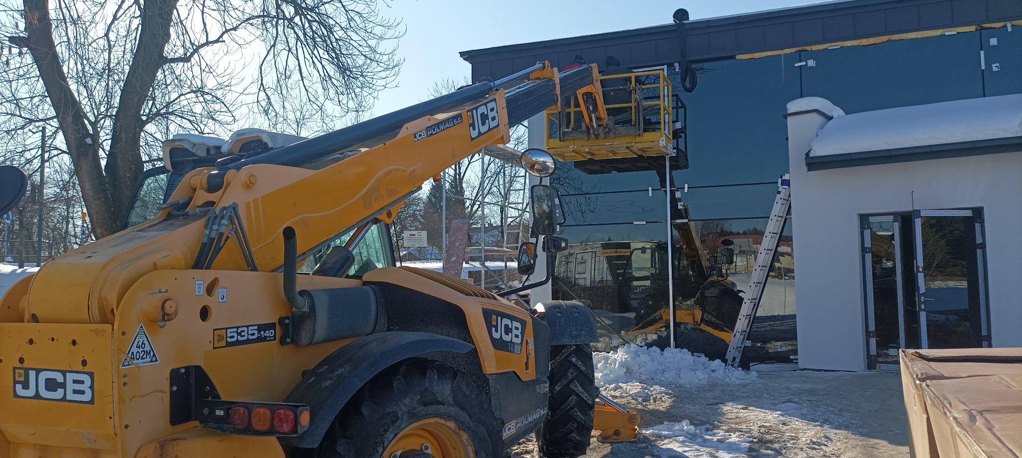 Montaż szklanej fasady budynku z użyciem podnośnika teleskopowego JCB na tle zimowego krajobrazu. Widoczna drabina i częściowo zamontowane okna.